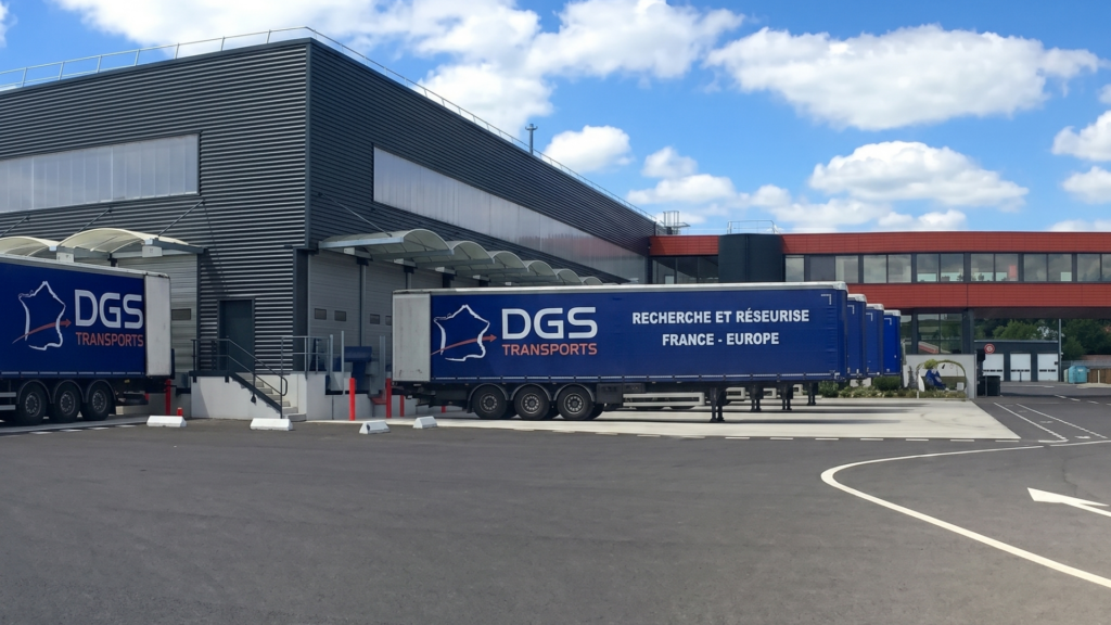 Several blue dgs transports semi-trailers parked at the loading docks of a large, modern grey warehouse under a bright blue sky with white clouds.
