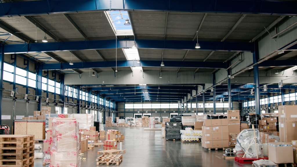 The interior of a large, brightly lit warehouse with a blue steel frame, showing various pallets of cardboard boxes and shrink-wrapped goods resting on a polished concrete floor.